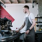 A young male barista wearing a grey apron operates an espresso machine in a modern café with industrial décor, preparing coffee drinks for customers.