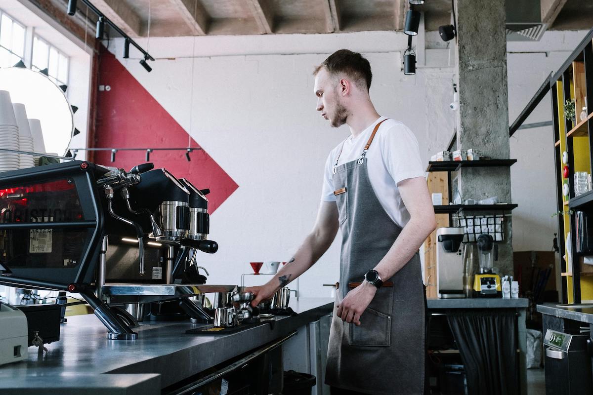 A young male barista wearing a grey apron operates an espresso machine in a modern café with industrial décor, preparing coffee drinks for customers.