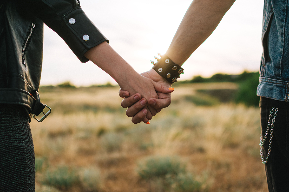 Two people in alternative-style clothing hold hands in a sunlit field, with warm light shining between their arms.