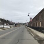 Hometown Heroes banners hang from lampposts along both sides of a Fitchburg street beside a long brick building with boarded windows. Snow remains along the sidewalks.